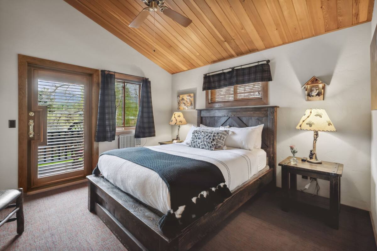Un dormitorio con techo de madera, una cama con colcha blanco y negro, dos lámparas de noche y una mesita con lámpara.