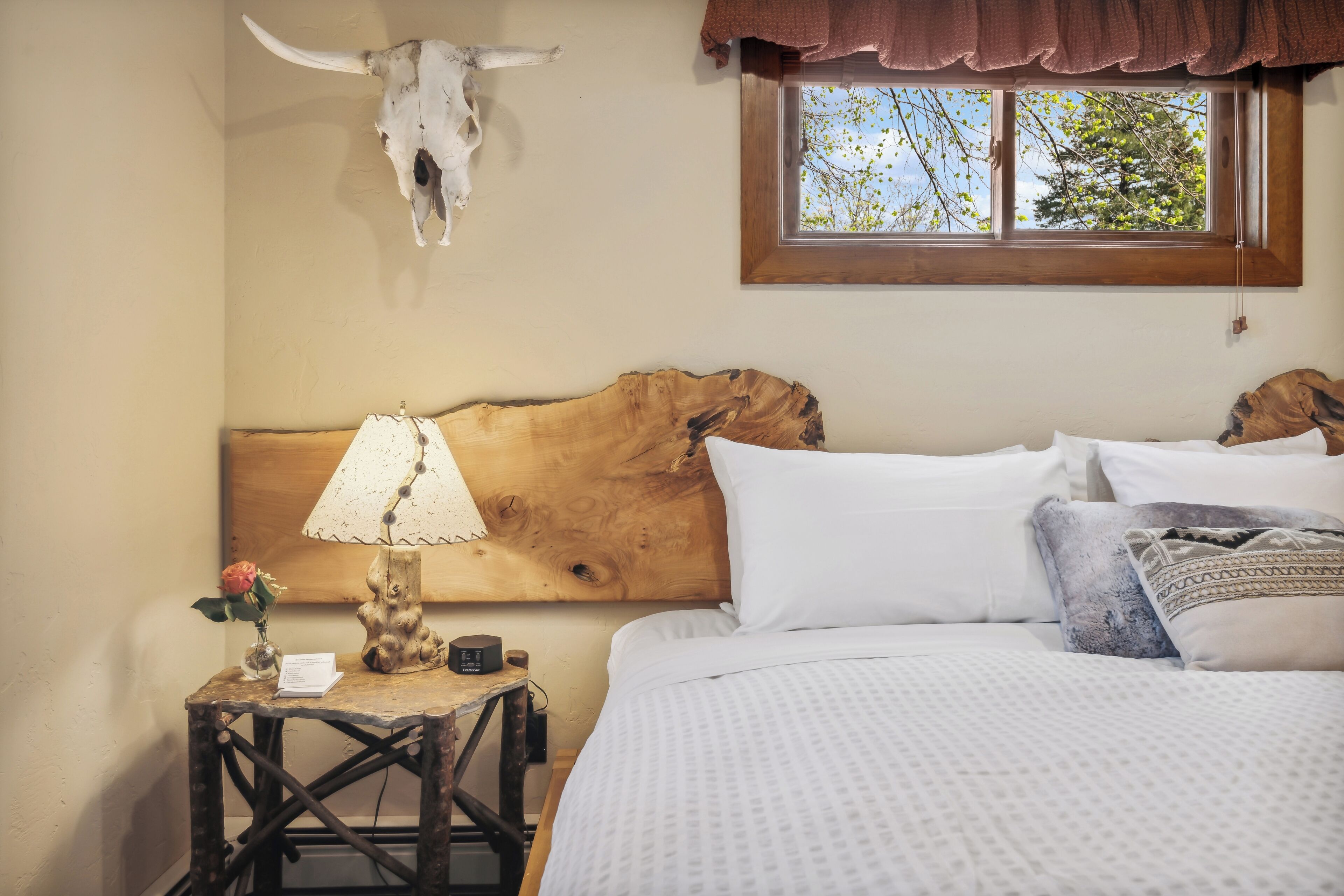 Un dormitorio con cabecera de madera, una mesita de noche con lámpara, un jarrón con flores y una ventana que da a un paisaje de árboles.