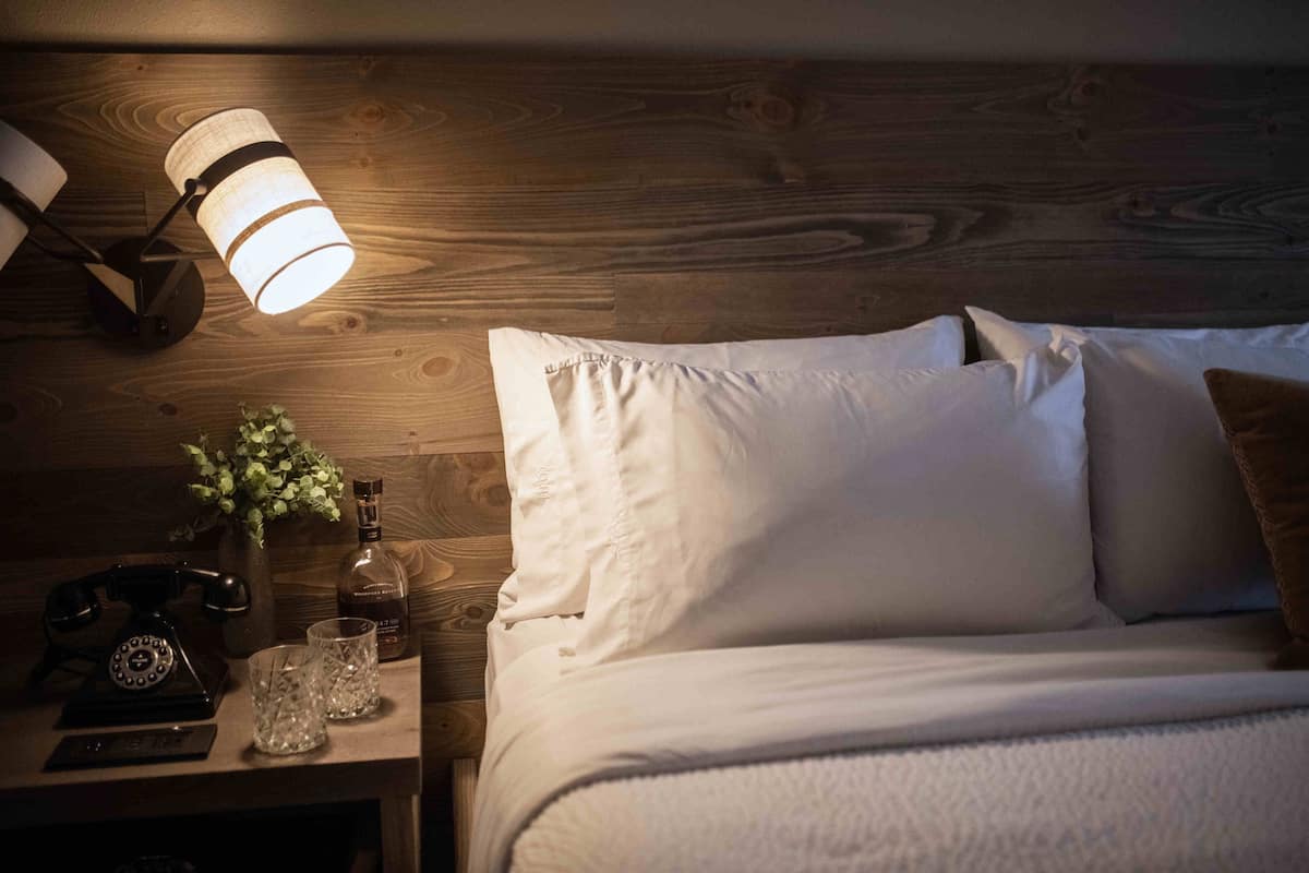 Ein ordentlich bezogenes Bett mit weißen Leinen, eine Nachttischlampe, ein altes Telefon und eine kleine Vase mit Blumen auf einem Holznachttisch.