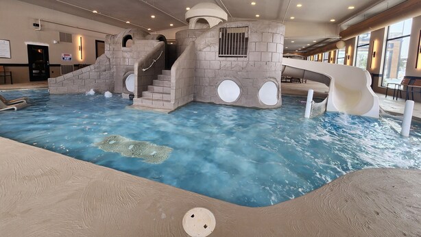 Ein großer Indoor-Wasserpark mit einem zentralen Pool, einer Rutsche und einer Treppe zu einer Plattform.