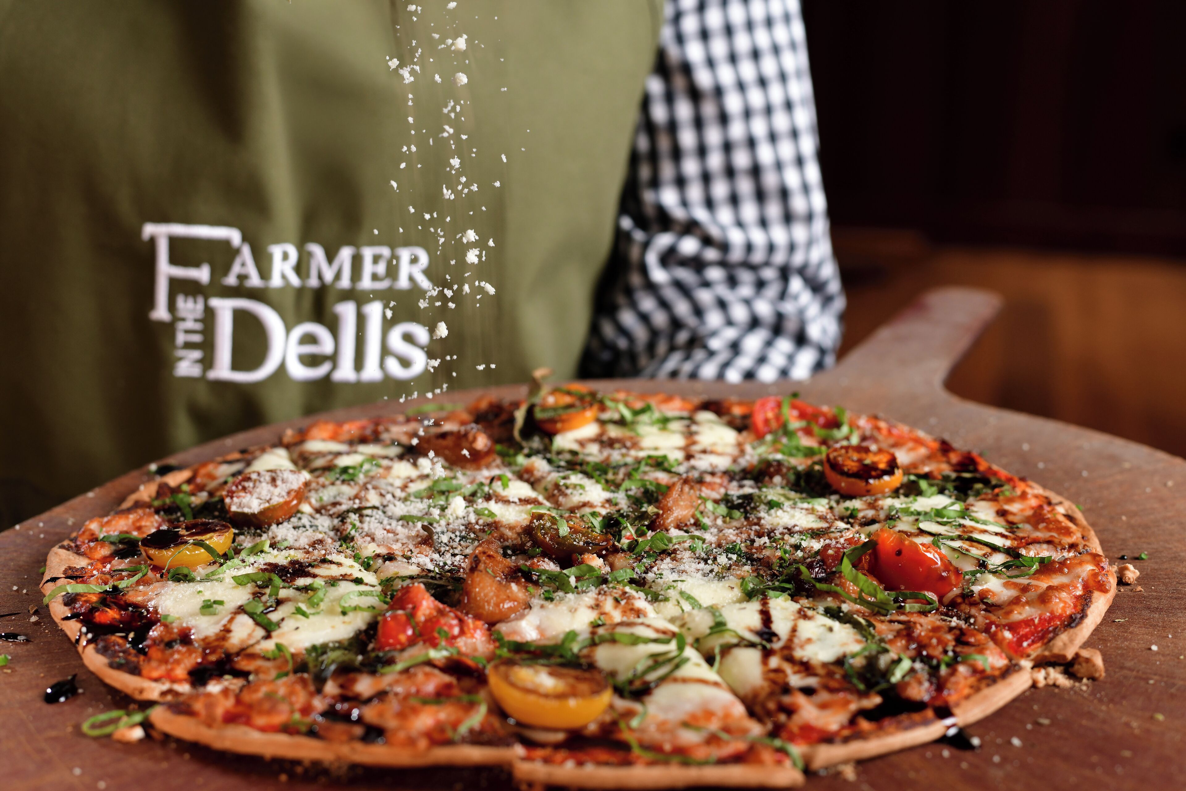 A pizza with various toppings, a wooden cutting board, and a person wearing a checkered apron.