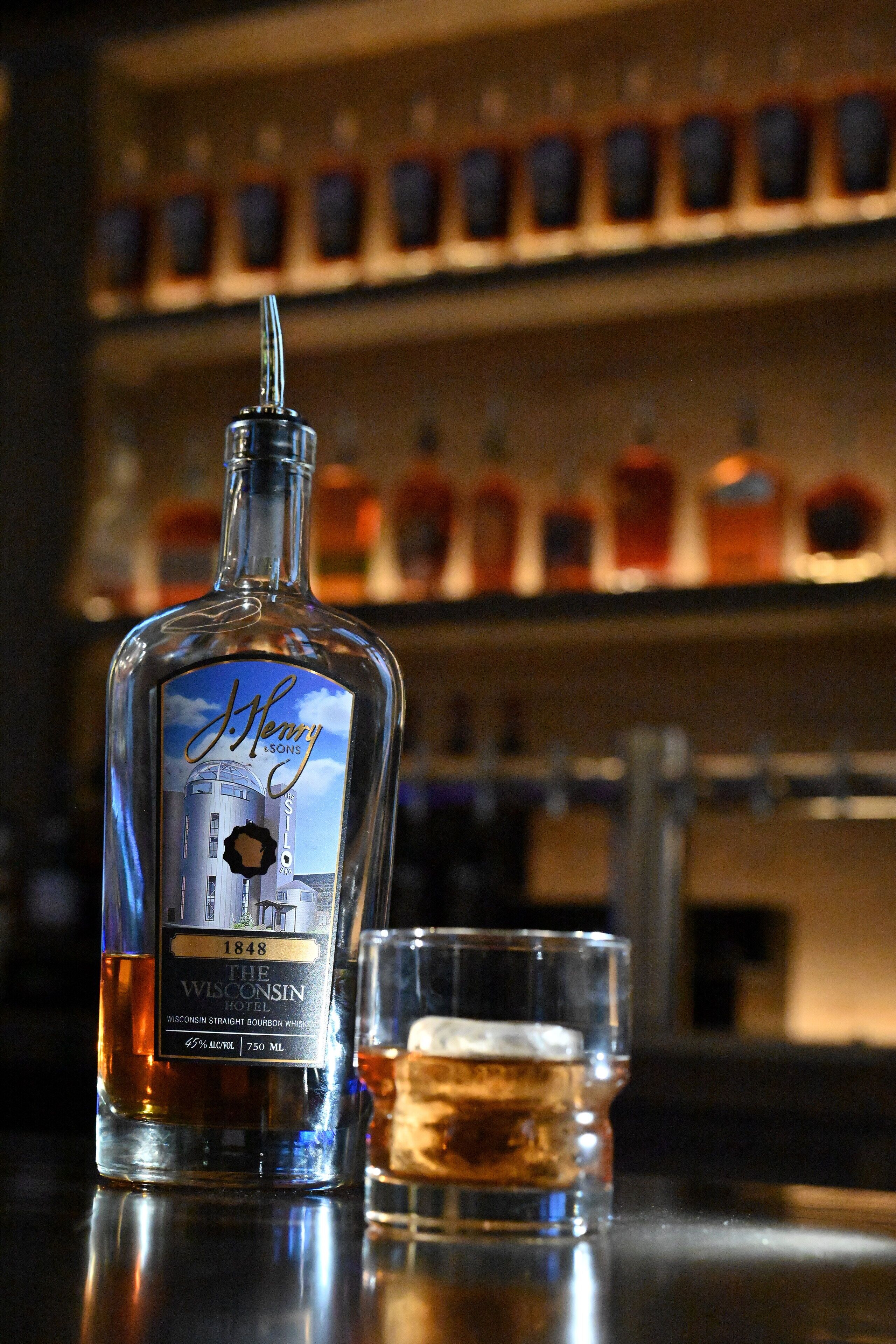 En flaske J. Henry's 1848 The Wisconsin Hotel whiskey med et glas whiskey ved siden af.