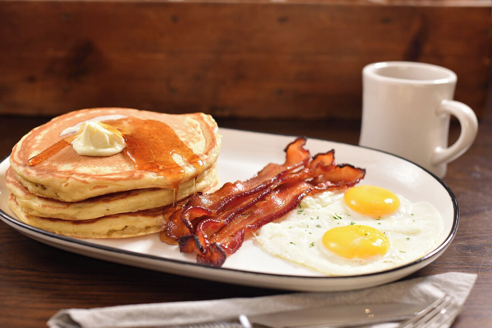 A plate of pancakes with syrup, bacon, and two fried eggs.
