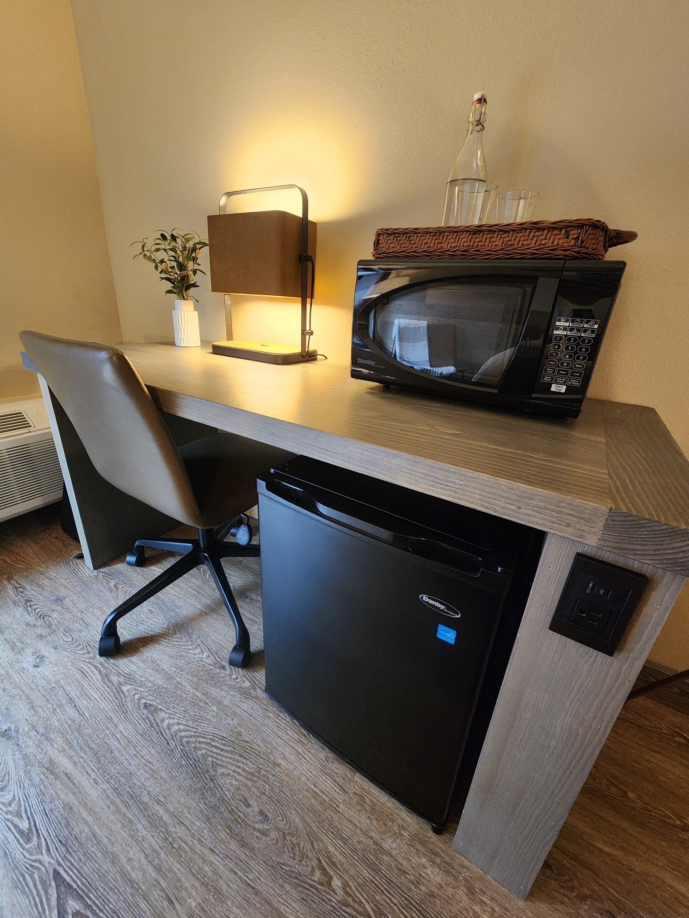 A compact kitchenette with a microwave, a small refrigerator, a chair, a table lamp, and a potted plant.