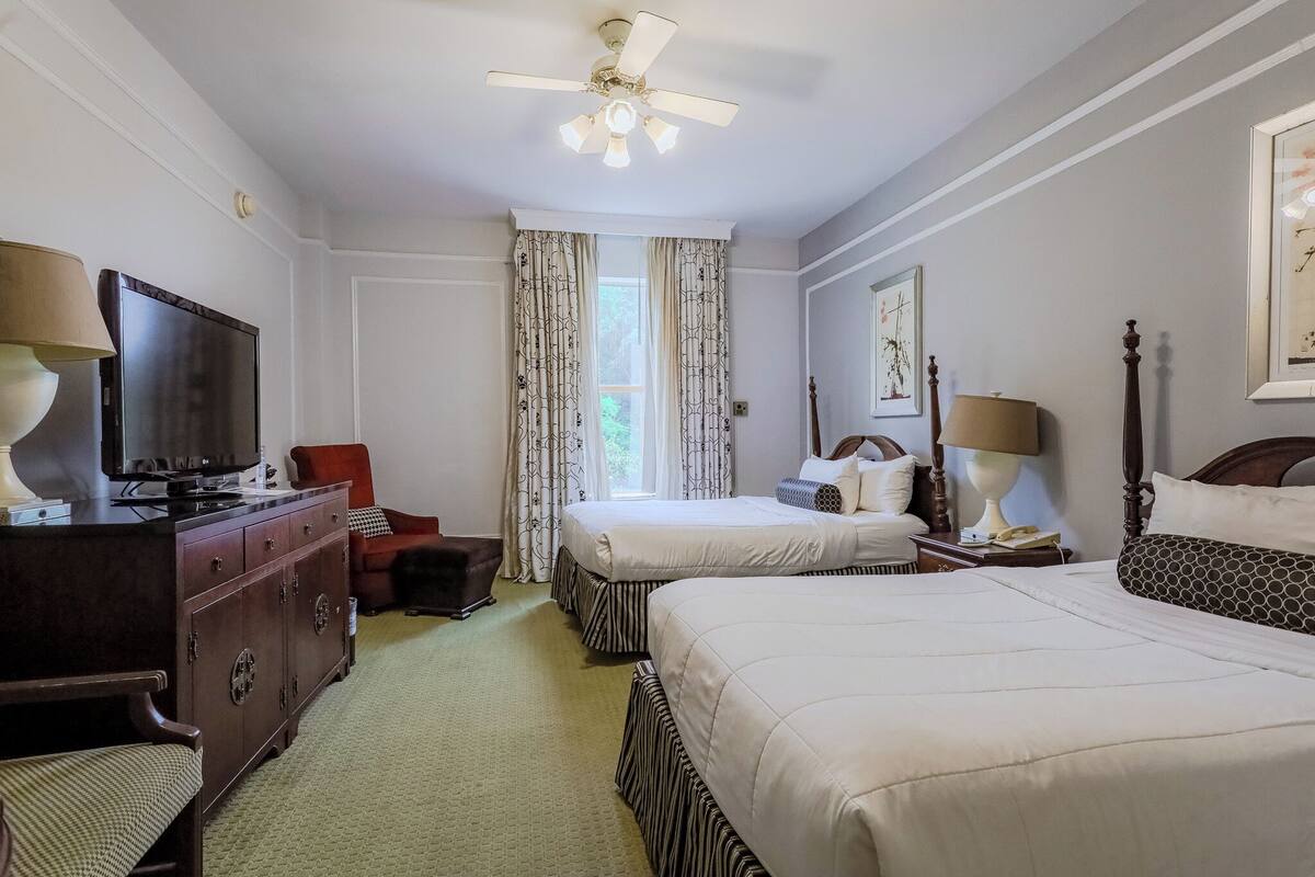 A hotel room with two beds, a television, a desk, a chair, a dresser, and a ceiling fan.