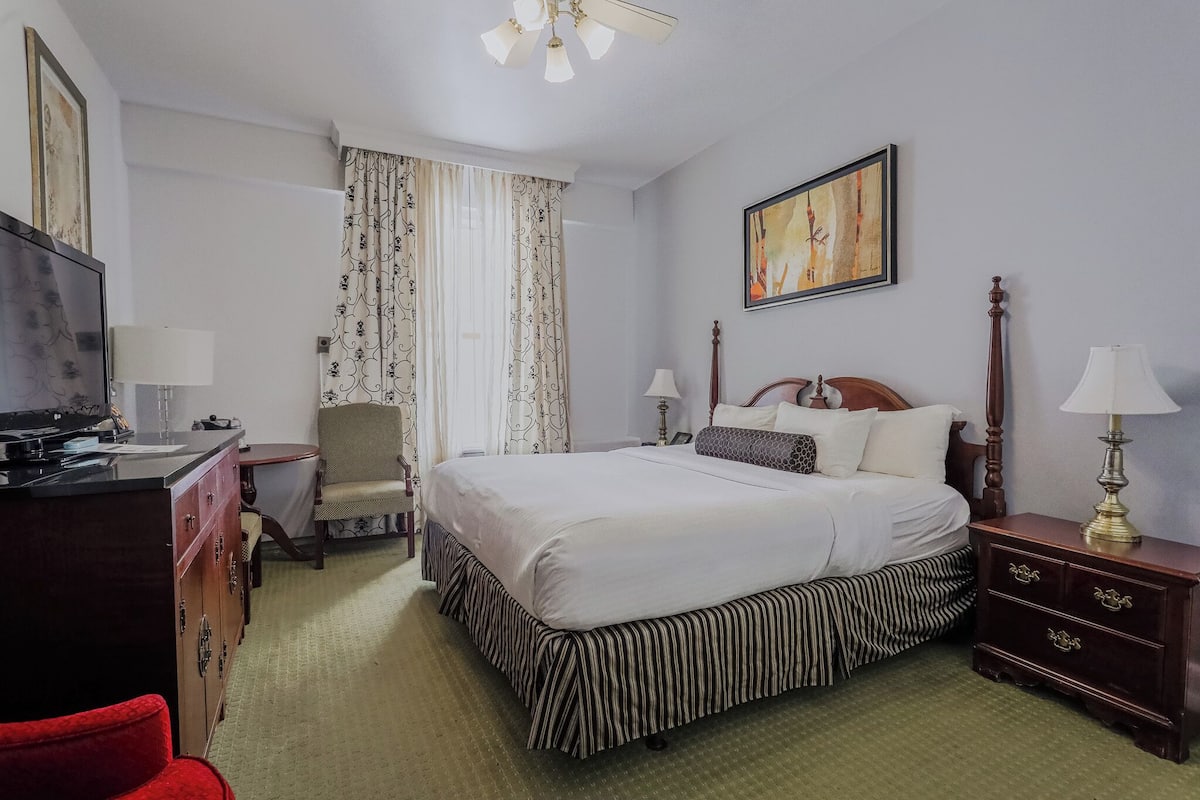 A hotel room with a bed, a desk, a chair, a television, and a painting on the wall.