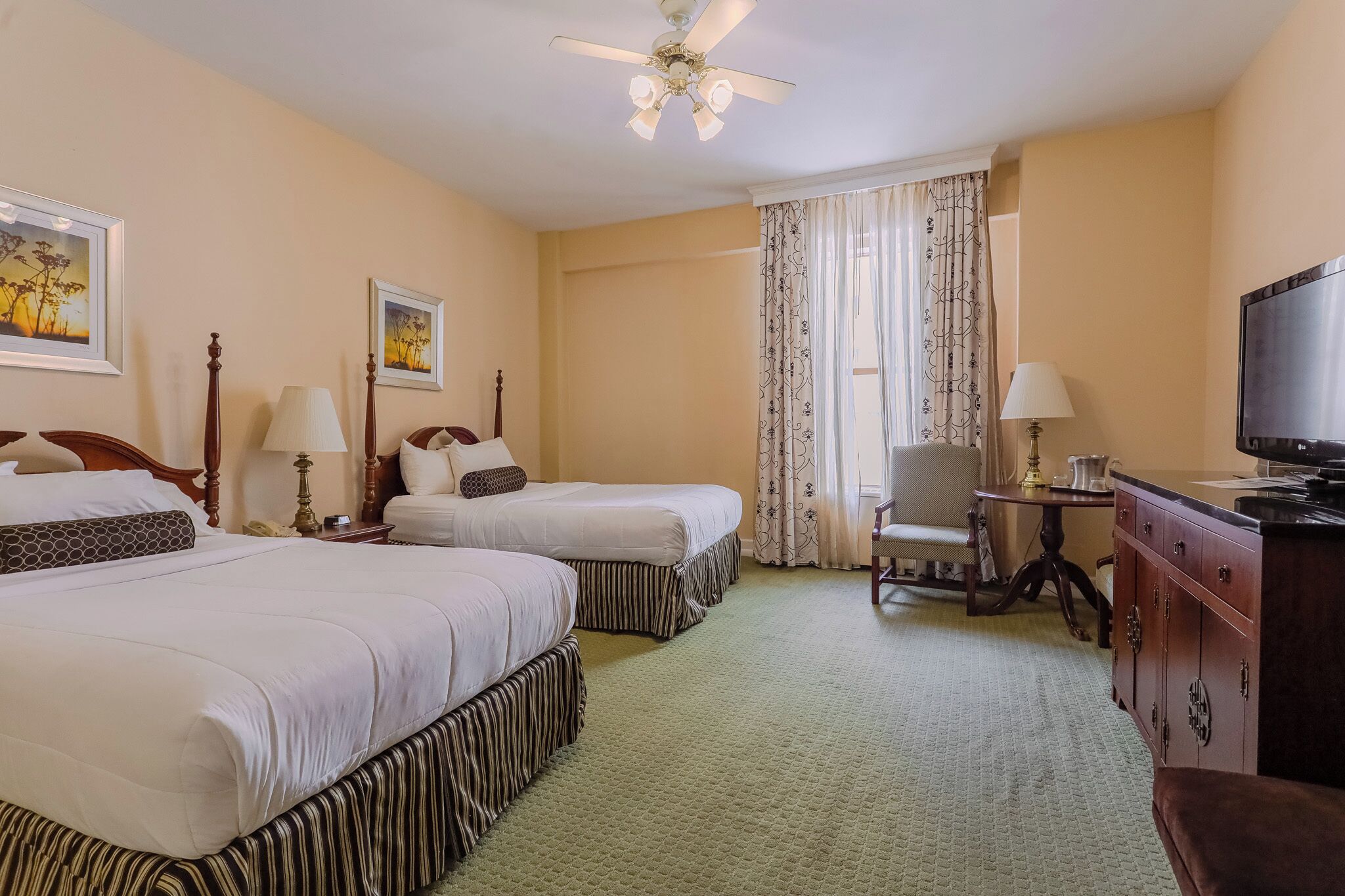 A hotel room with two beds, a TV, a desk, a chair, and a ceiling fan.