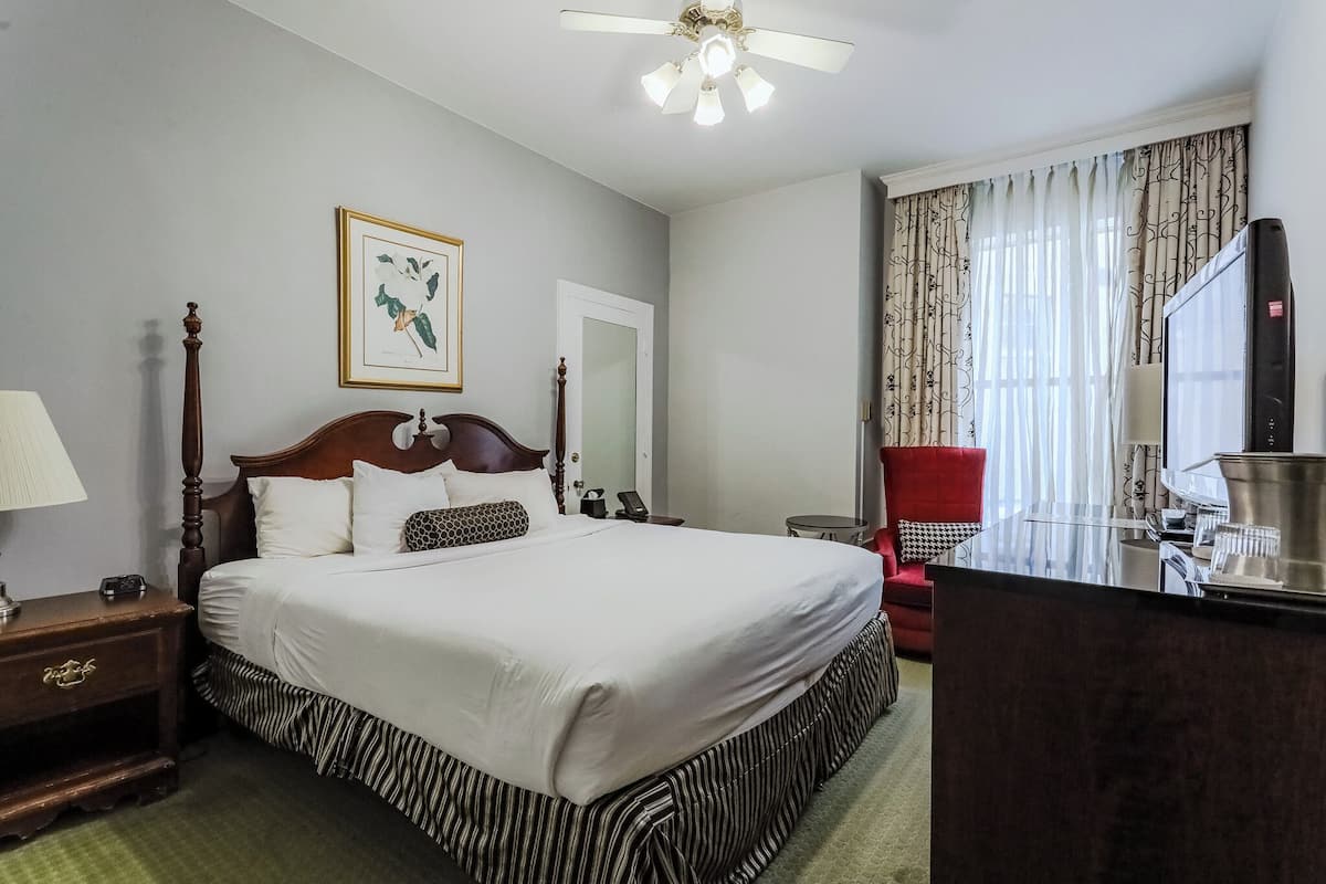 A hotel room with a large bed, a wooden headboard, a ceiling fan, a framed botanical print, a red chair, a desk with a mirror, and a television.