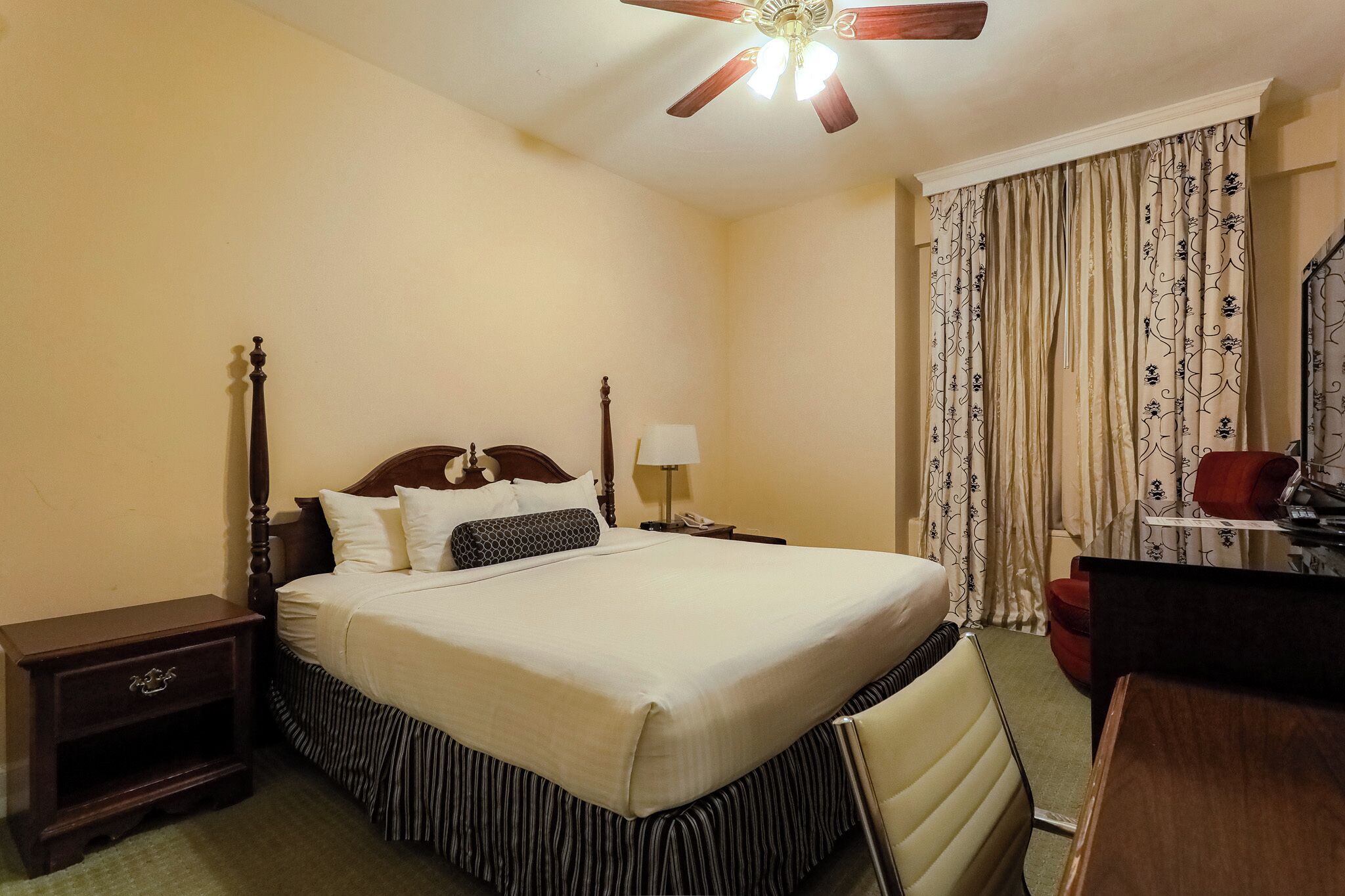 A hotel room with a bed, a nightstand, a desk, a chair, a ceiling fan, and curtains.