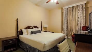 A hotel room with a bed, a nightstand, a desk, a chair, a ceiling fan, and curtains.
