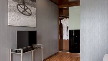 Una habitación de hotel moderna con un televisor de pantalla plana grande sobre un soporte, suelo de madera, armario con batas y una pared con textura y un diseño similar a un grafiti.