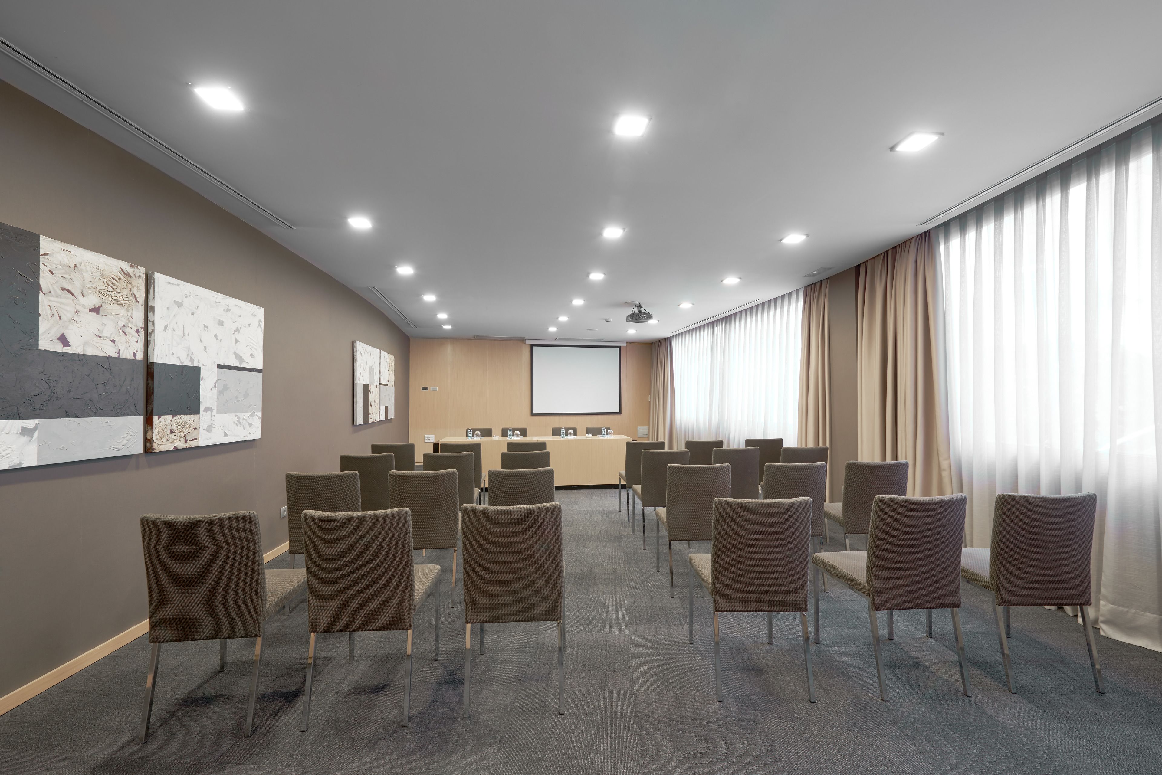 Un salón de conferencias con filas de sillas, una pantalla de proyector y un amplio ventanal con cortinas.