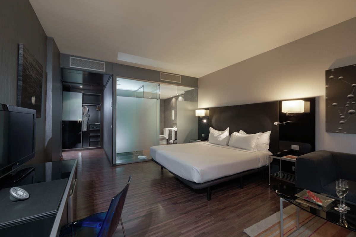 A modern hotel room with a large bed, a desk with a computer, a TV, and a glass partition separating the room from a bathroom.