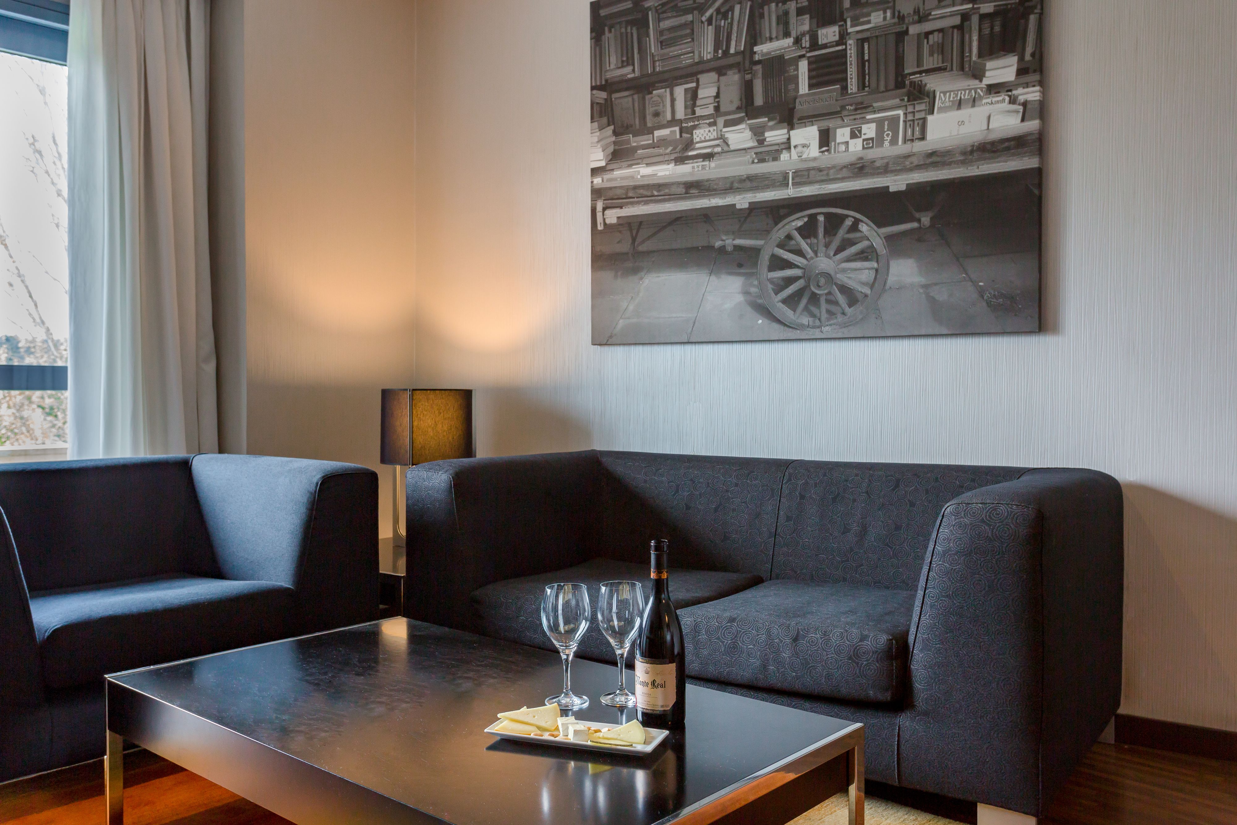 A modern living room with a dark grey sofa, a wooden coffee table with a bottle of wine and glasses, and a bookshelf with books and a framed picture.