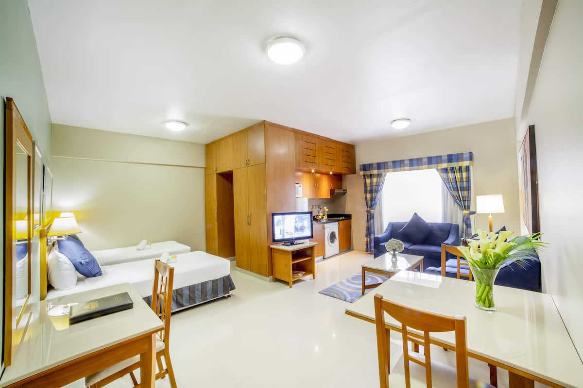 A compact hotel room with a bed, a desk with a chair, a small kitchenette, a sofa, and a dining table.