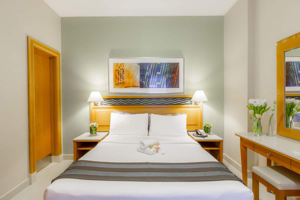 A hotel room with a bed, two bedside tables, a nightstand, a desk, a chair, a mirror, and a painting on the wall.