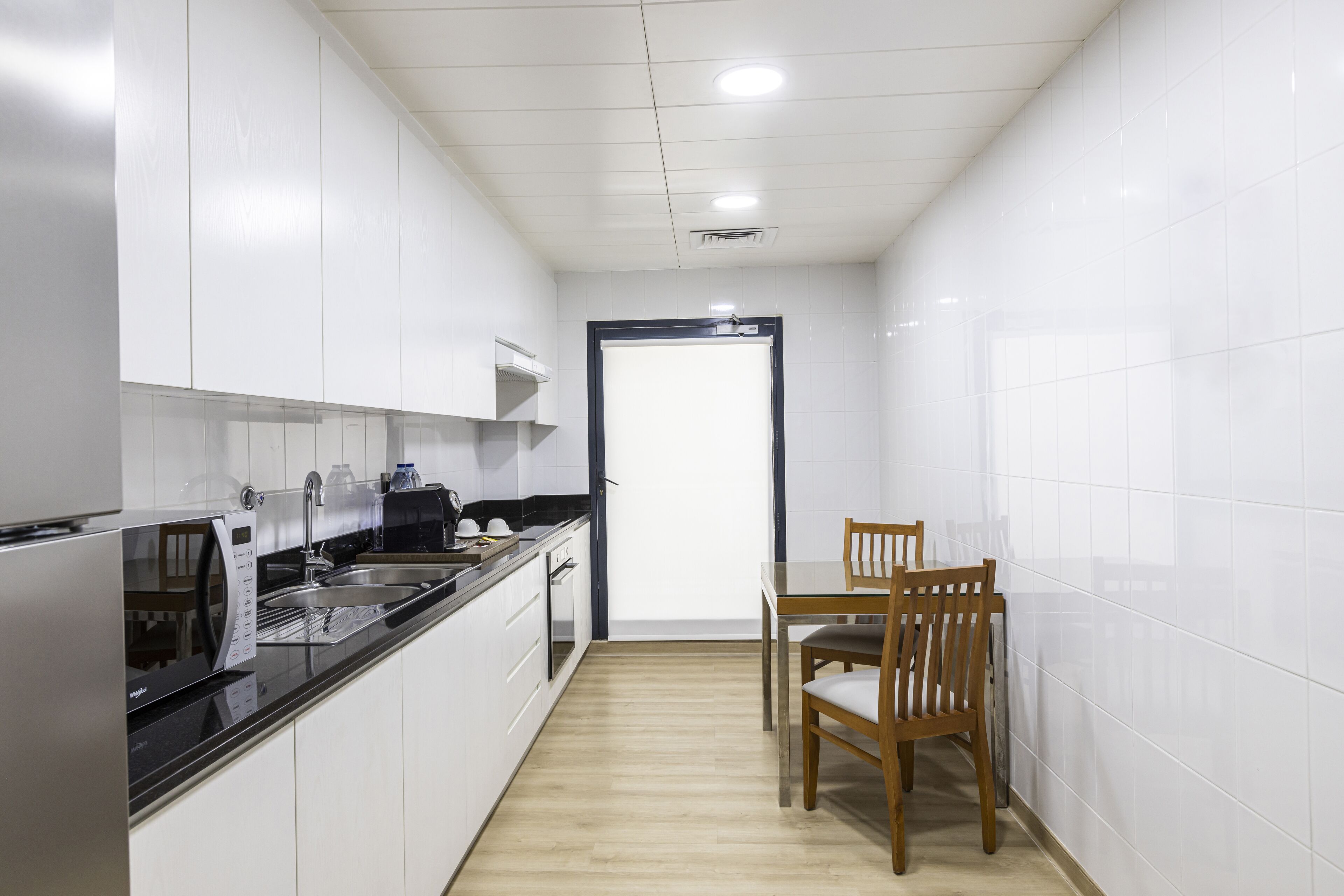 Une cuisine moderne avec des meubles blancs, un plan de travail noir, une table à manger en bois et une porte donnant sur une autre pièce.