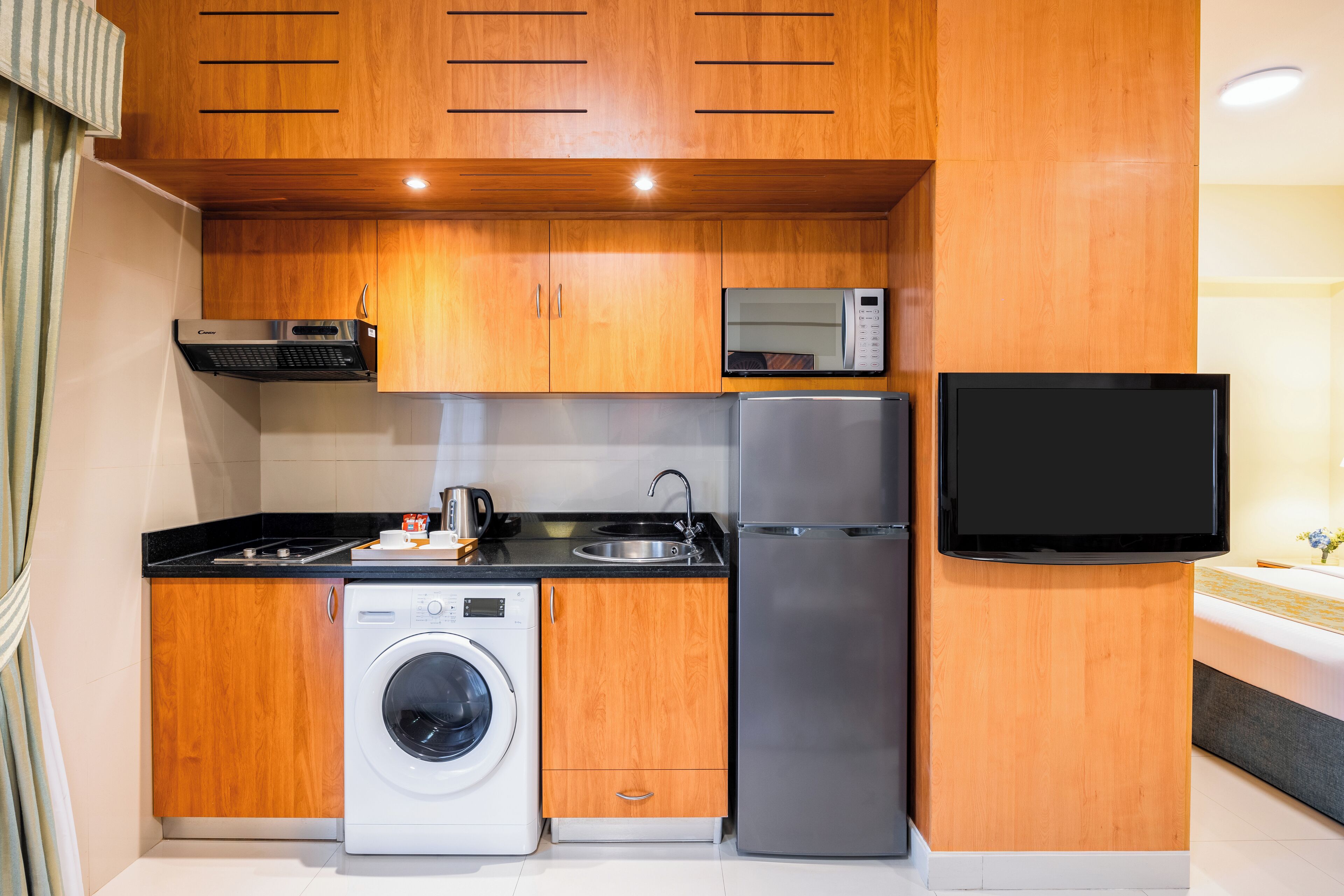 A compact kitchen with wooden cabinets, a washing machine, a refrigerator, a microwave, and a flat-screen TV.