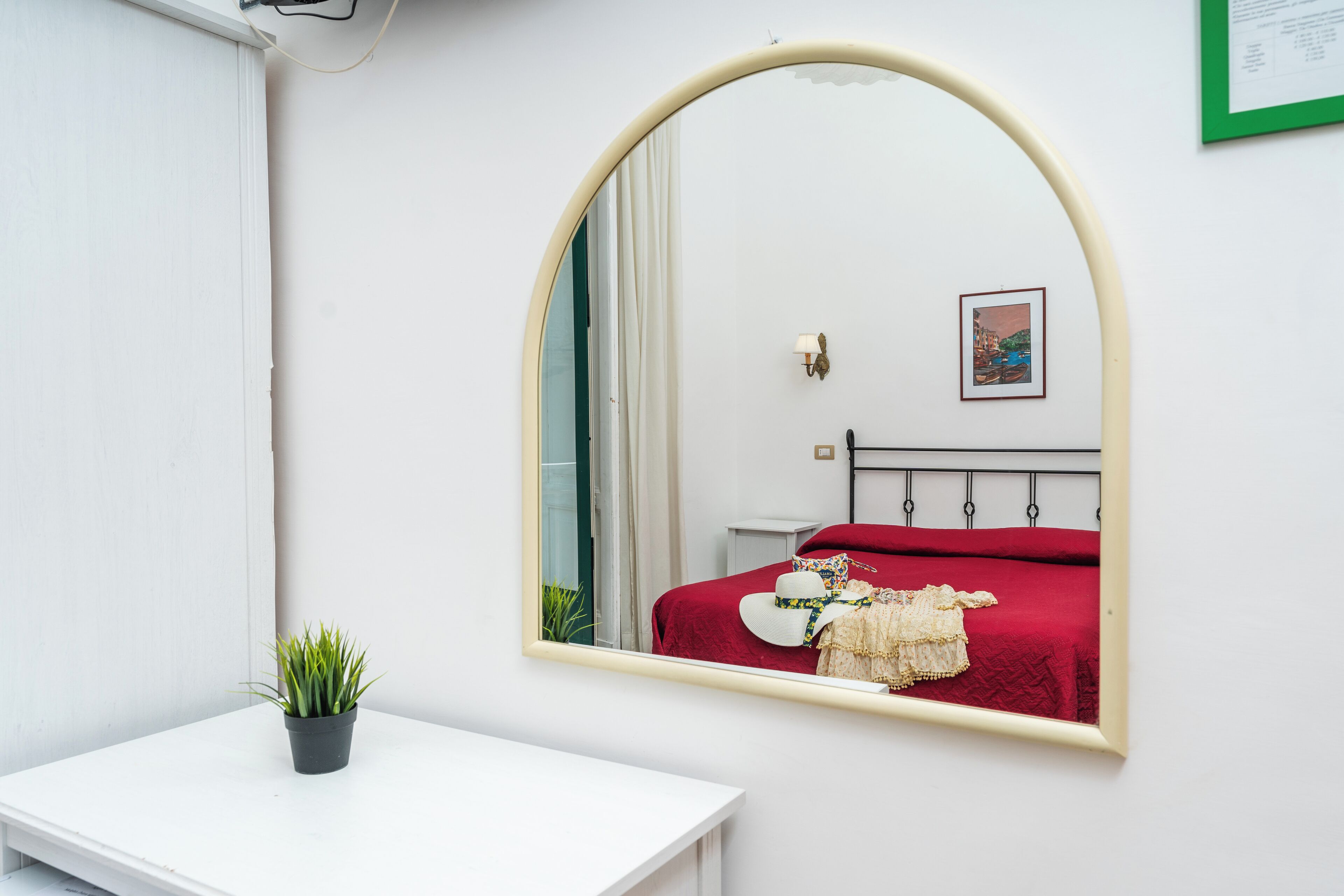 A bedroom with a white wall, a large mirror, a bed with red bedding, a small potted plant, and a framed picture.