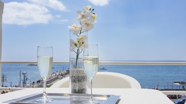 Una mesa con dos copas de vino blanco y un florero con flores blancas, con vista a una playa y cielo despejado.