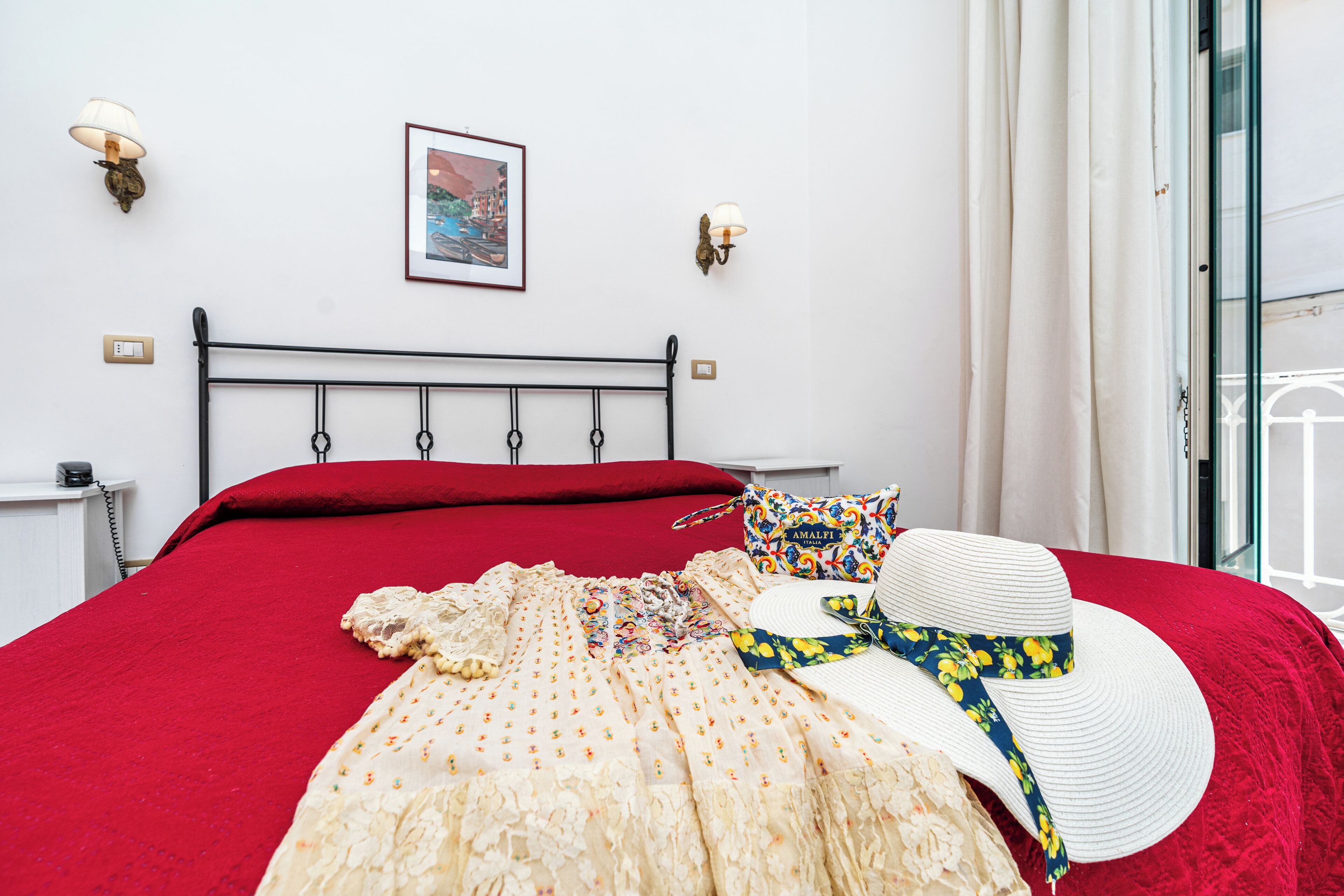 Un lit bien fait, recouvert d’une couverture rouge, avec une robe à motifs floraux et un chapeau de soleil posés dessus.