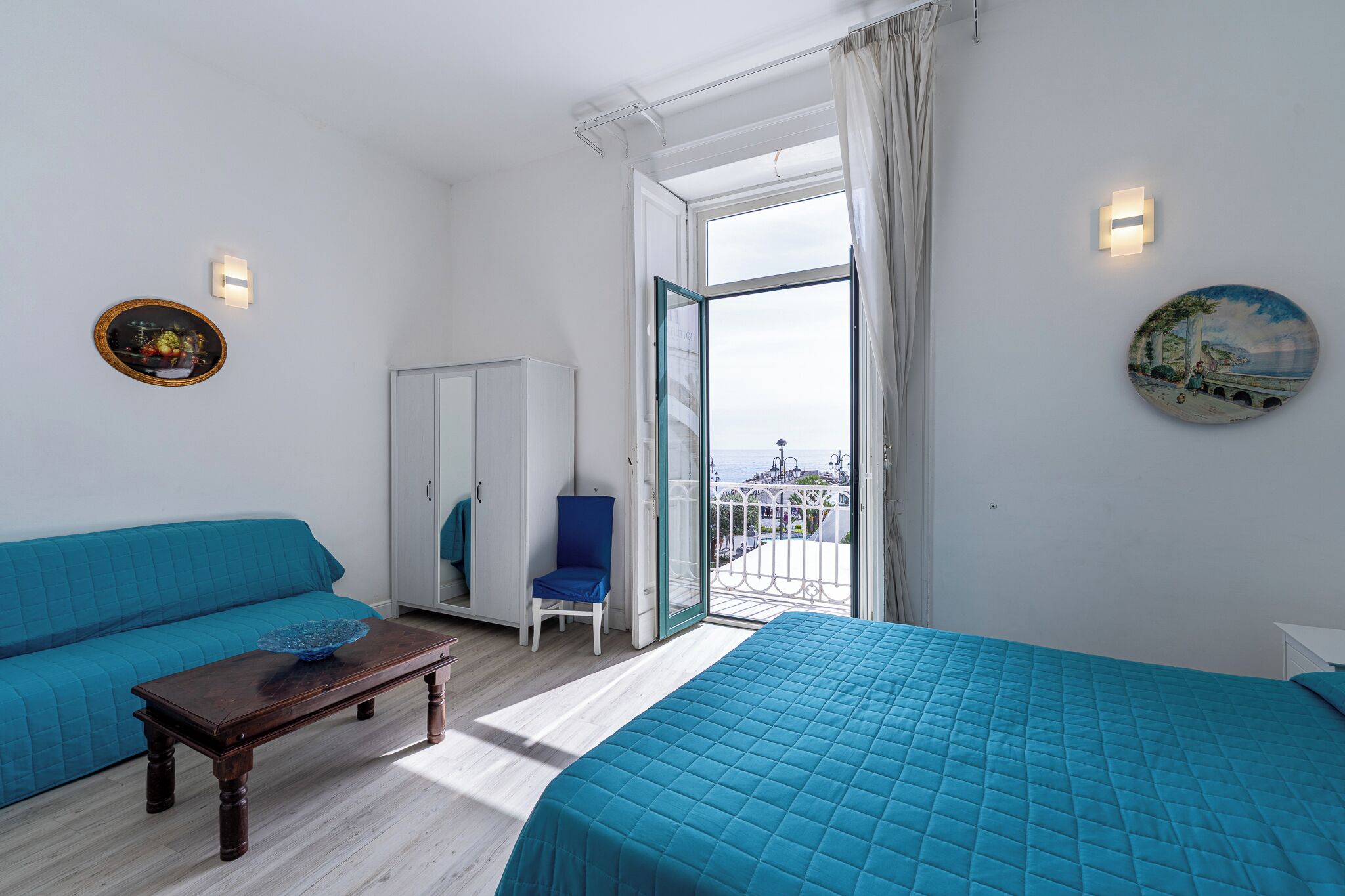 A room with a bed, a blue sofa, a chair, a table, a wardrobe, and a balcony with a view.