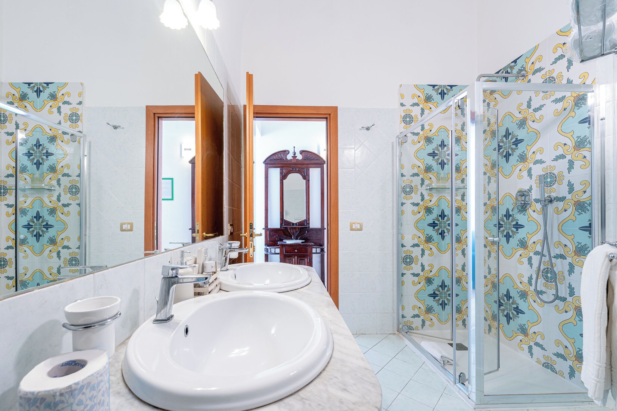 A bathroom with two sinks, a toilet, a mirror, and a shower with patterned tiles.