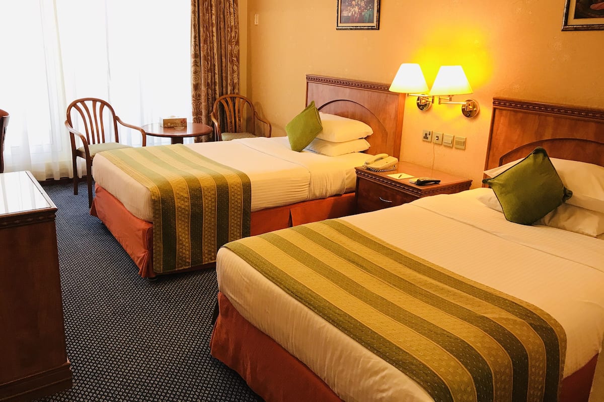 Habitación de hotel con dos camas, cabecero de madera, mesita de noche, lámpara y una silla.
