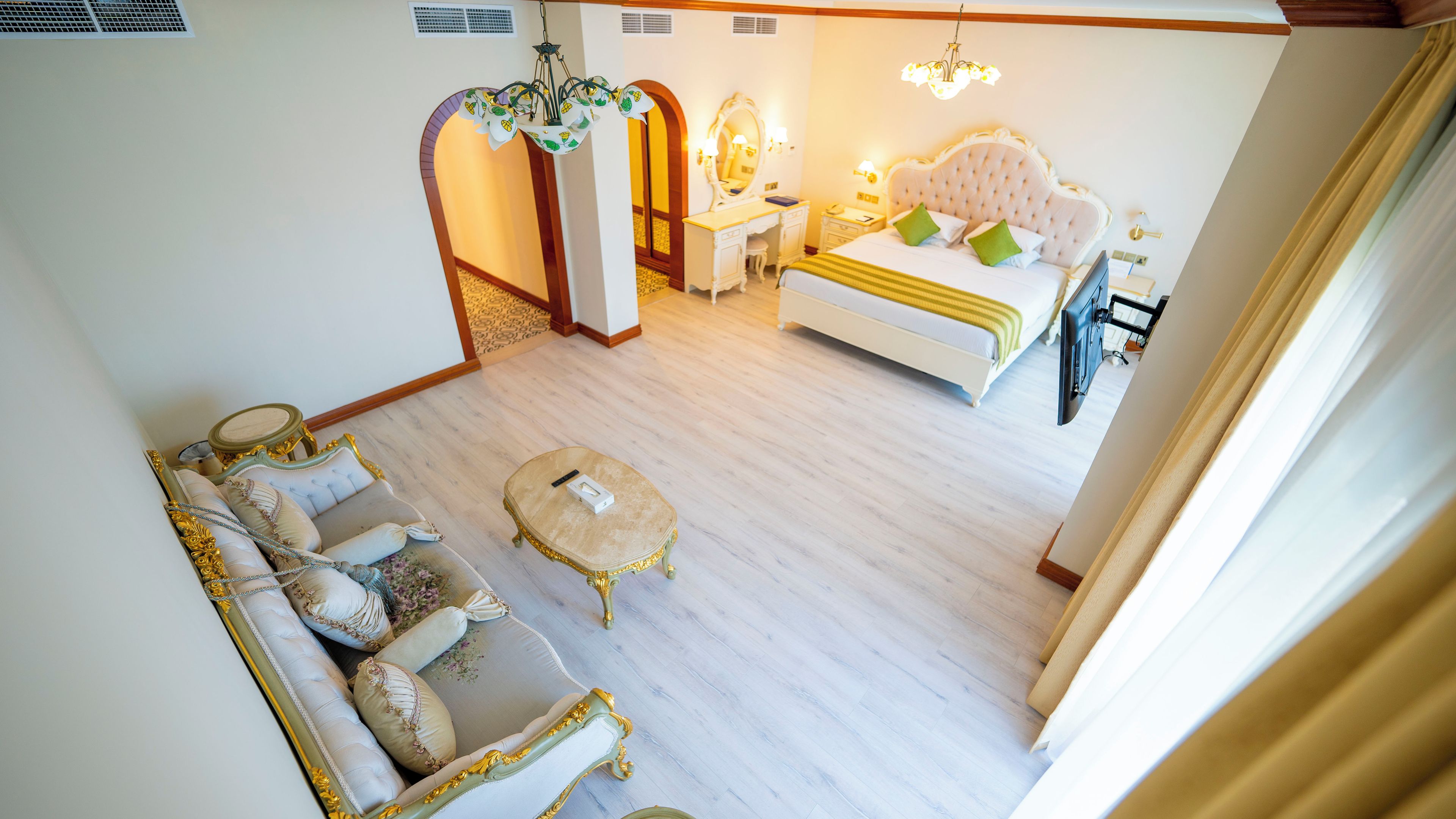 A hotel room with a bed, a sitting area with a sofa and a small table, a dressing area with a mirror and vanity, and a chandelier.