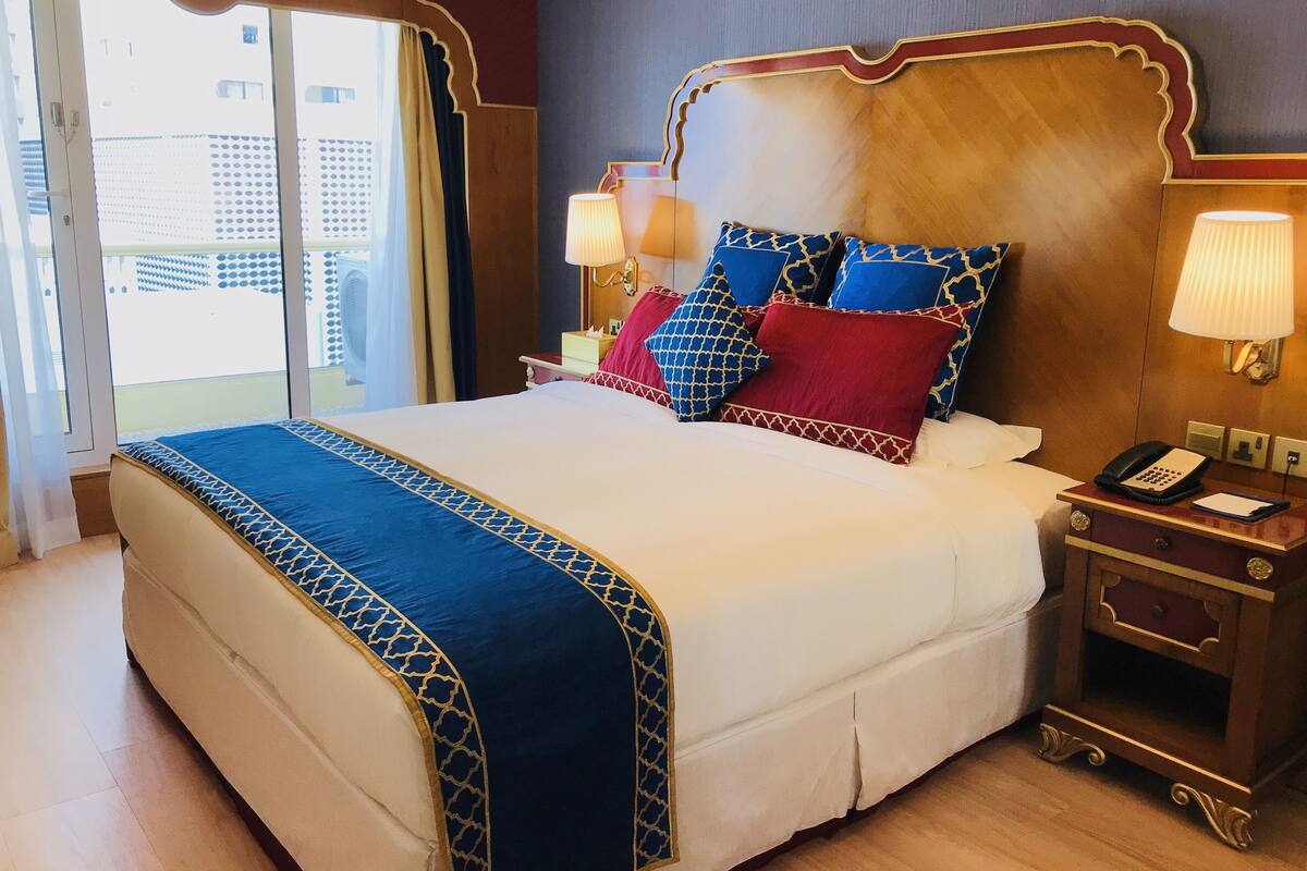 Una cama grande con una colcha estampada, un cabecero de madera y varias almohadas. Hay una mesita de noche con una lámpara y un teléfono.