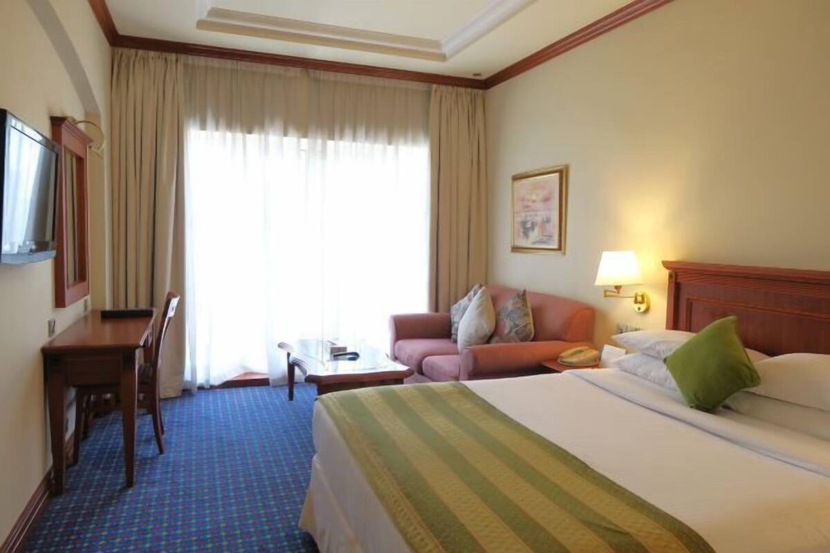 Una habitación de hotel con cama, escritorio, silla, mesita, sofá y ventana con cortinas.