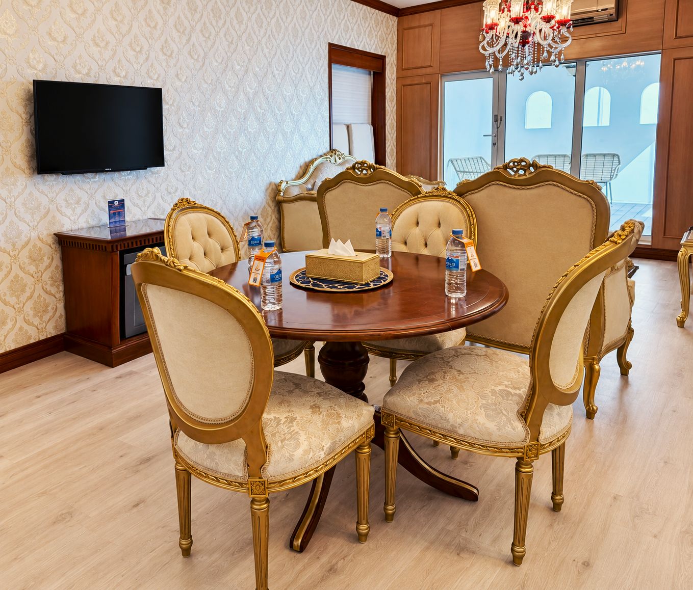 Een eetruimte met een ronde tafel, zes beklede stoelen met gouden accenten, een houten buffetkast en een flatscreen televisie aan de muur.