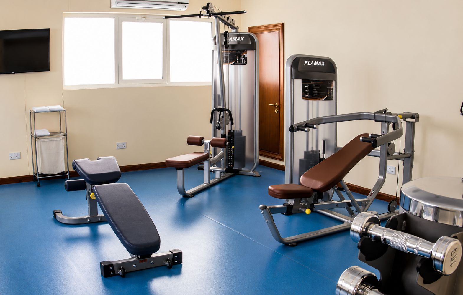 Un gimnasio con equipo de ejercicio, una banca, un televisor montado en la pared y un ventanal.