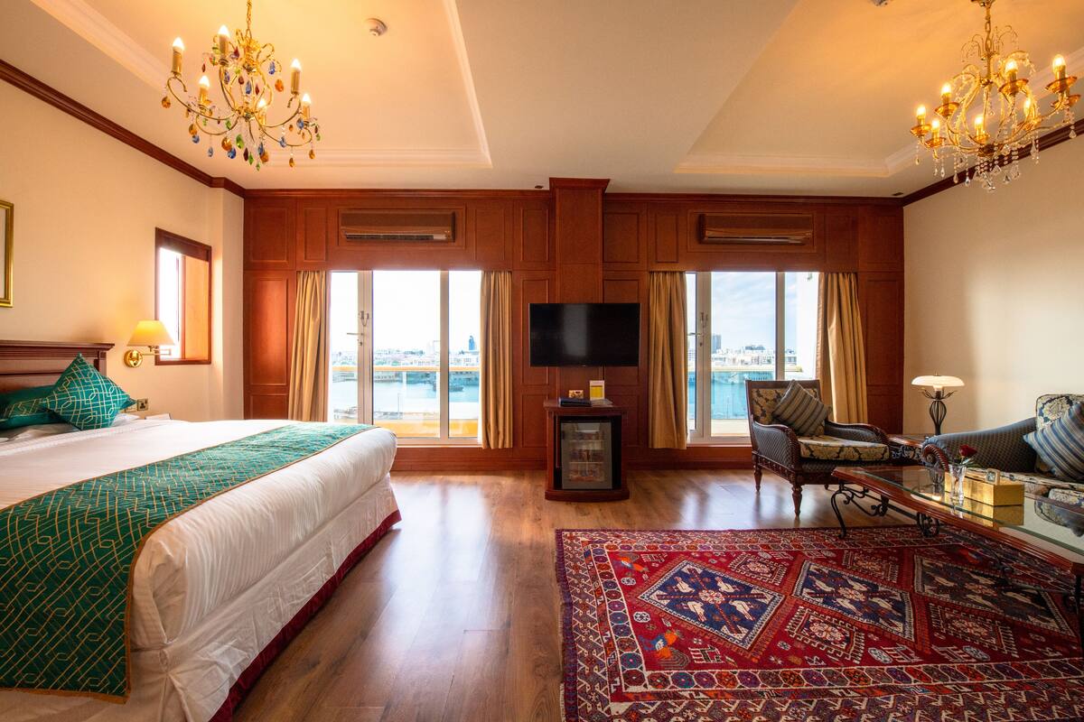 Habitación de hotel con una cama grande, televisión, zona de estar con sofá y sillón, y vista de la ciudad a través de amplias ventanas.