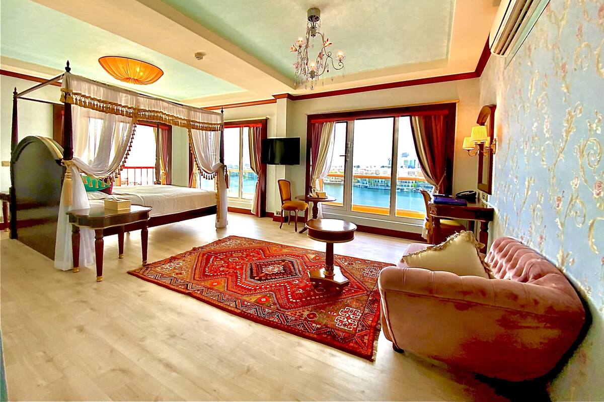 Una habitación amplia con cama con dosel, zona de estar con sofá y un amplio ventanal con vistas a la ciudad.