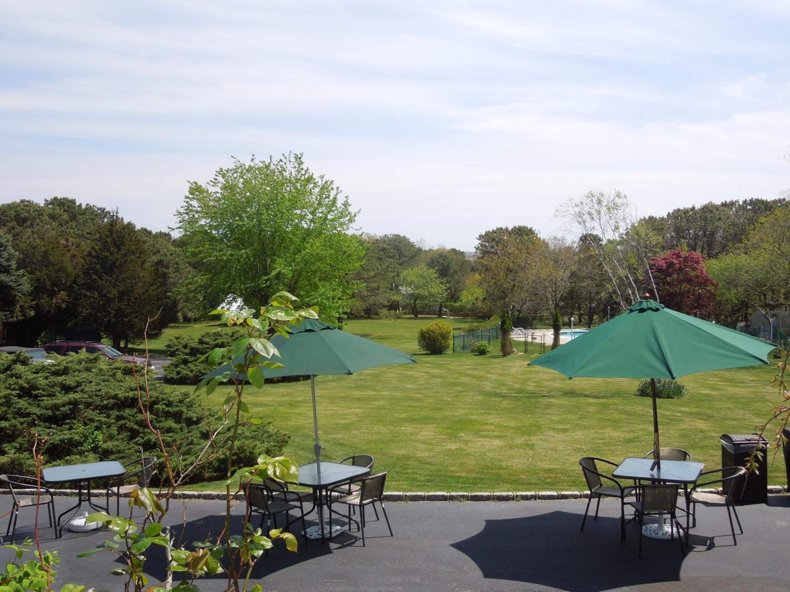 An outdoor patio area with tables and chairs under green umbrellas, surrounded by a grassy lawn and trees.