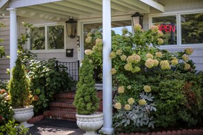 A front porch with a white column, a potted plant, and a hedge with pink flowers.