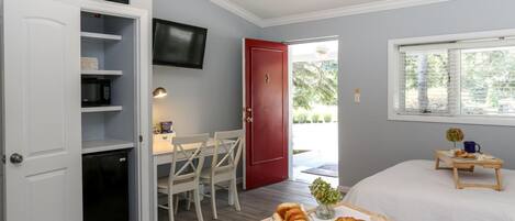 A compact room with a bed, a small table with breakfast items, a TV mounted on the wall, and a red door leading outside.