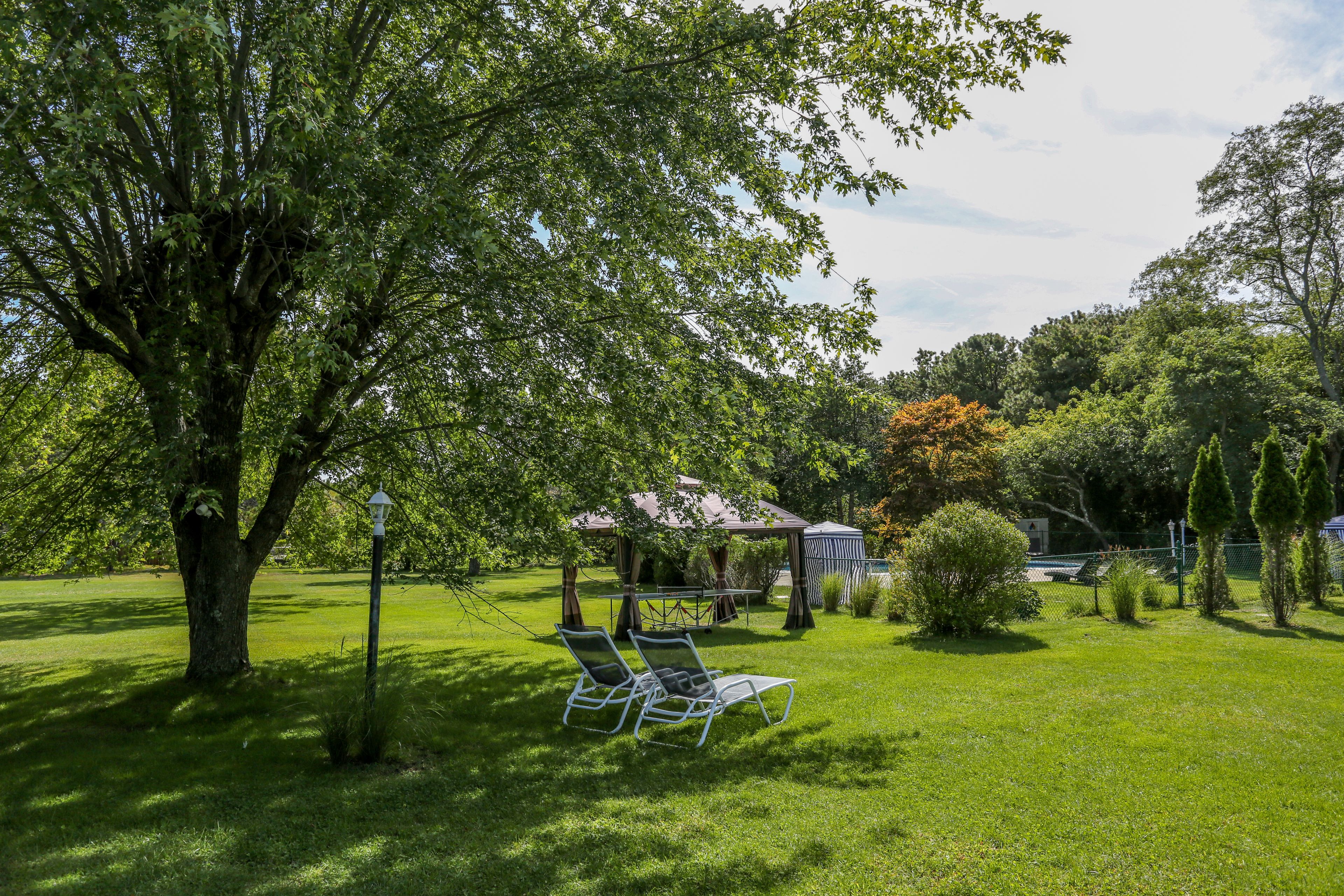 A grassy outdoor area with two metal chairs under a tree, a gazebo, and a small building in the background.