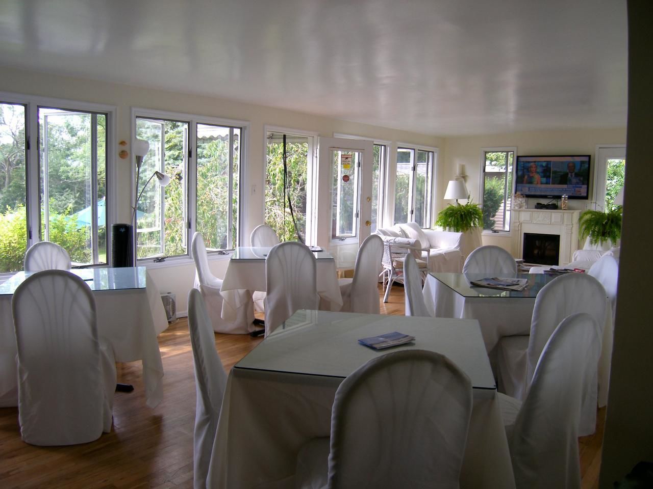 Un vaste espace dédié à la salle à manger, avec des chaises et des tables blanches, de grandes fenêtres et un téléviseur fixé au mur.
