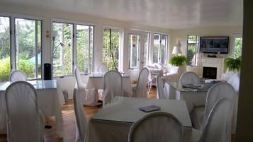 Un vaste espace dédié à la salle à manger, avec des chaises et des tables blanches, de grandes fenêtres et un téléviseur fixé au mur.