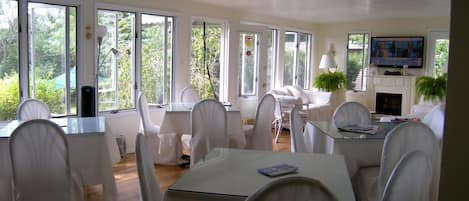 A spacious dining area with white chairs and tables, large windows, and a television on the wall.