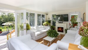 A spacious living room with a white sofa, a coffee table, and a fireplace. There are large windows offering a view of the outdoors. - Hamlet Inn (Southampton)