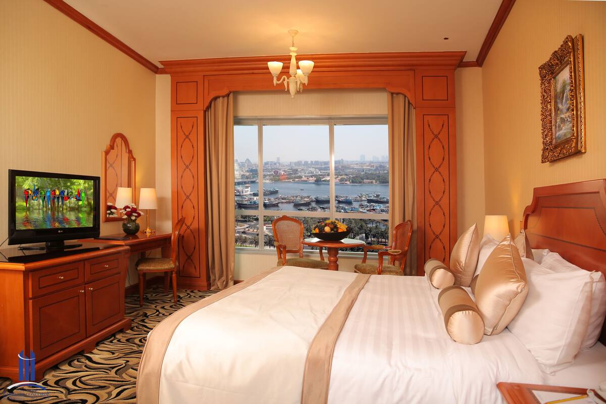 A hotel room with a large bed, a television, a dining table with chairs, and a view of a cityscape through the window.