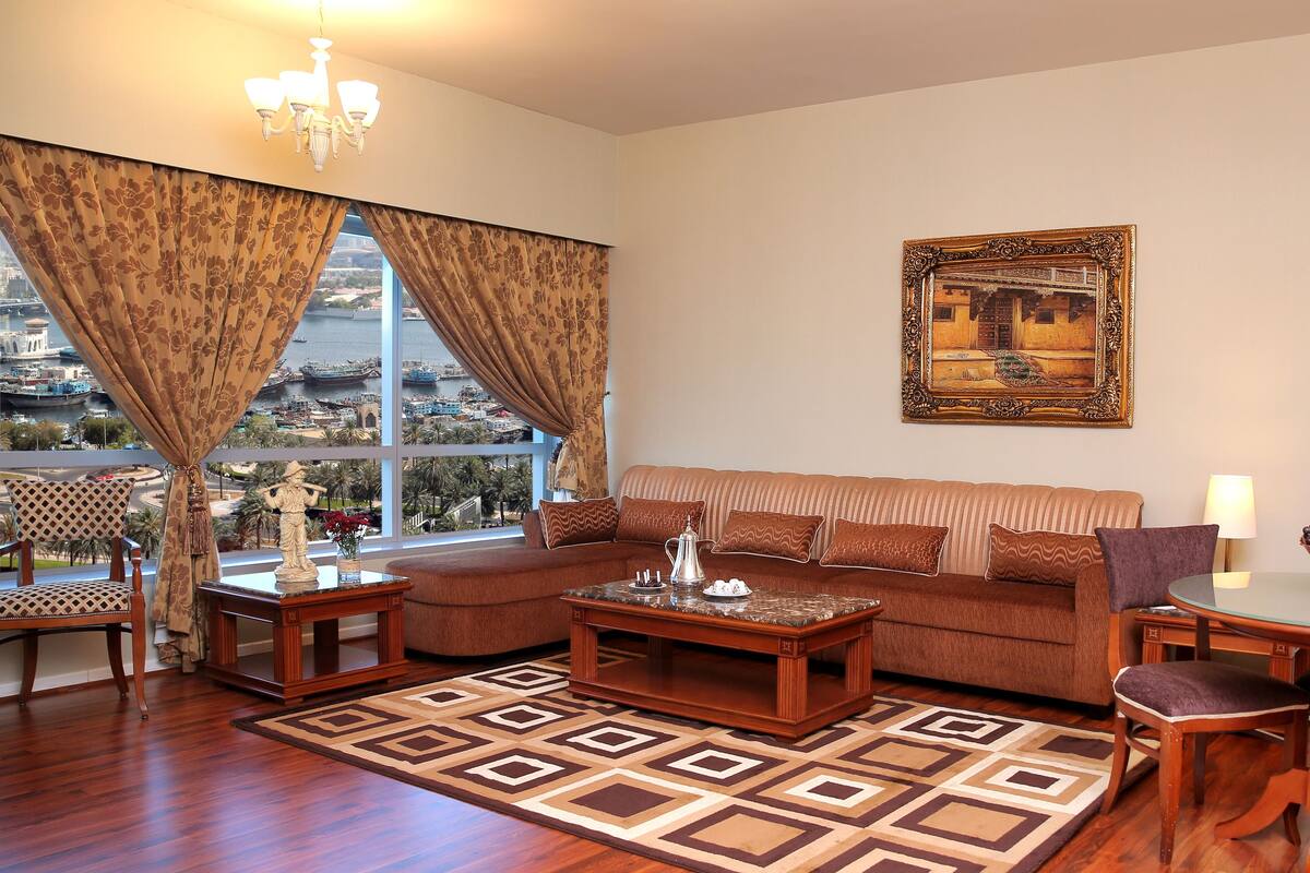 A living room with a sofa, coffee table, and chairs, a dining table with chairs, a chandelier, and a large window with curtains.