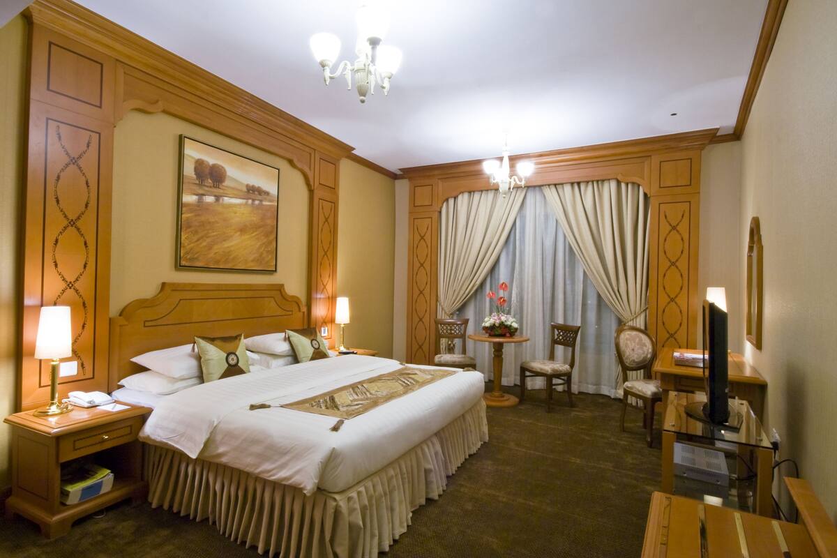 A hotel room with a large bed, two bedside tables with lamps, a desk with a chair, a television, and a window with curtains.