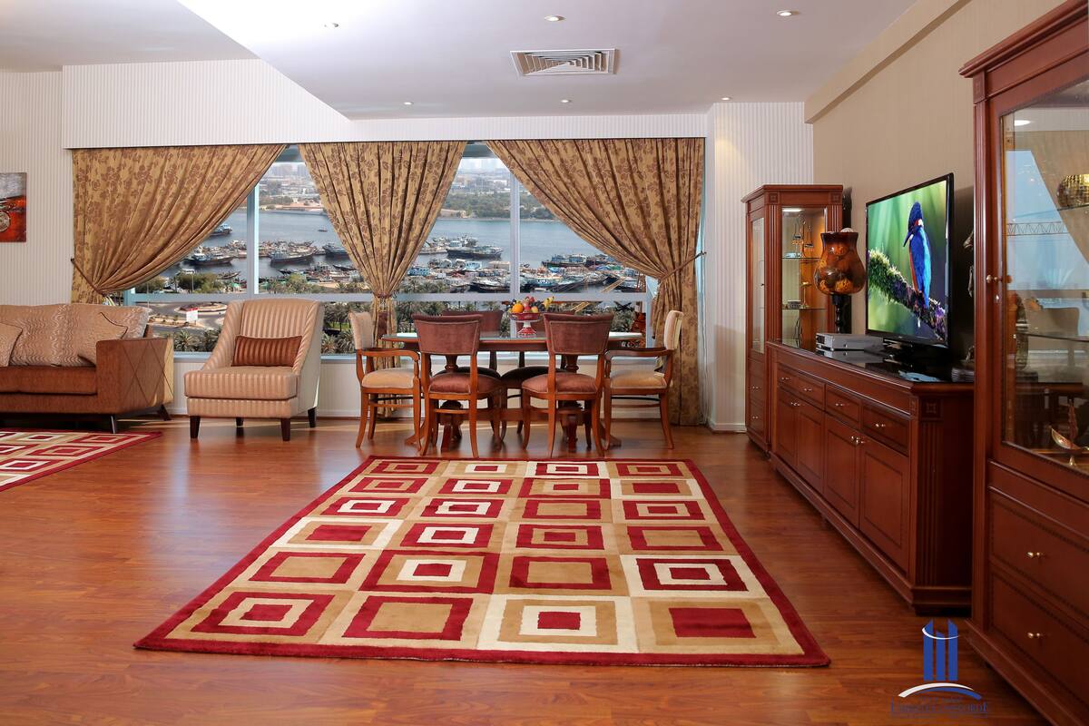 A spacious living room with a large window offering a view of a harbor, a dining area with a table and chairs, and a television on a wooden cabinet.