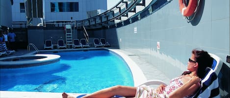 A pool area with a person lying on a lounge chair, a lifebuoy, and a building with a modern design in the background.