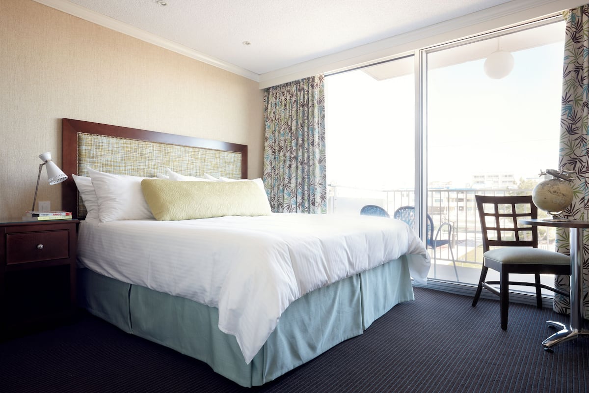 Habitación de hotel con una cama grande, un escritorio con silla, una mesita de noche y vistas al exterior desde un balcón.