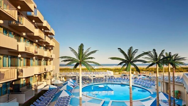 A hotel pool area with sun loungers, palm trees, and a clear view of the ocean. - Oceanfront Pan American Hotel (Wildwood Crest)
